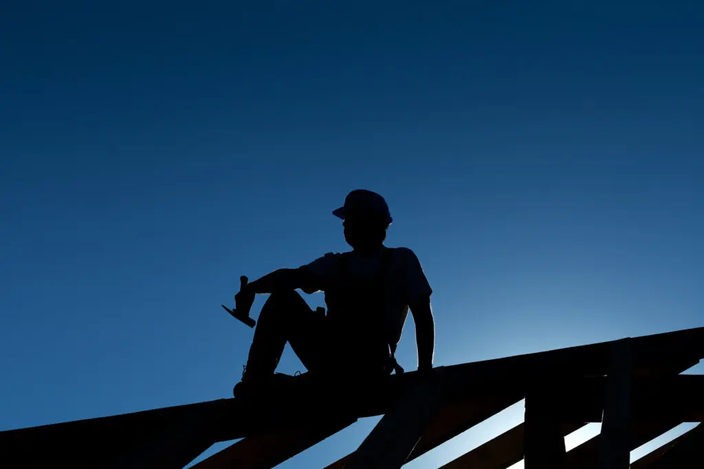Silhouette of construction worker sitting on roof framing holding roofing tool at sunset
